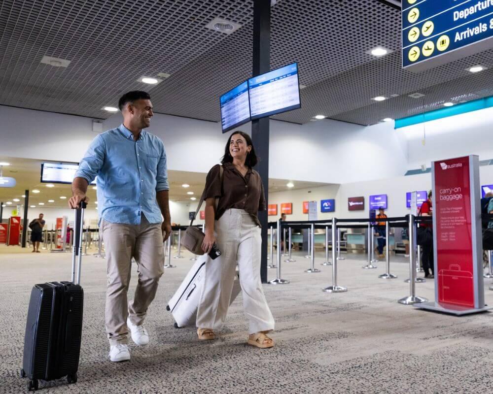 Passengers with luggage walk through a busy Townsville Airport check-in area, with multiple airline counters and digital flight information screens visible in the background. 