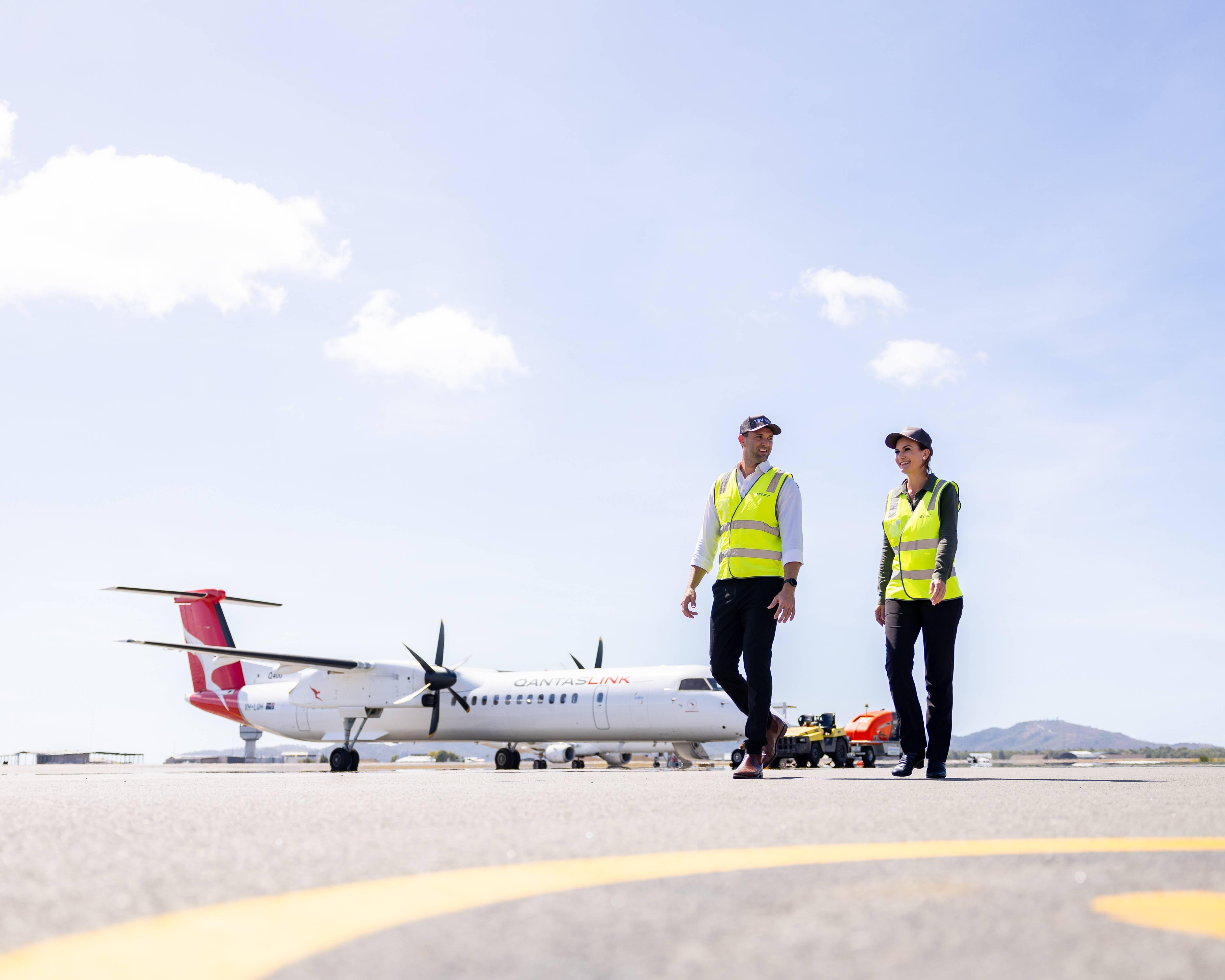 Two airport ground crew members in high-visibility vests walk across the tarmac near a QantasLink aircraft, set against a backdrop of mountains and a clear sky. 