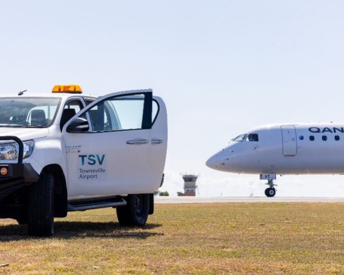 Townsville Airport operations vehicle near runway with QantasLink aircraft taxiing and control tower in background.