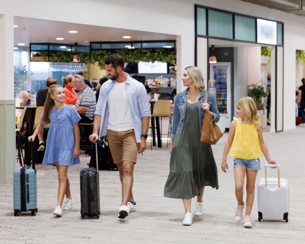 A family of four walking through the Townsville airport terminal with luggage, passing a dining area in the background.