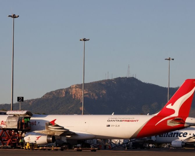 Qantas aircraft at Townsville Airport with cargo being loaded, surrounded by ground crew and equipment, with a mountain and communication towers in the background.