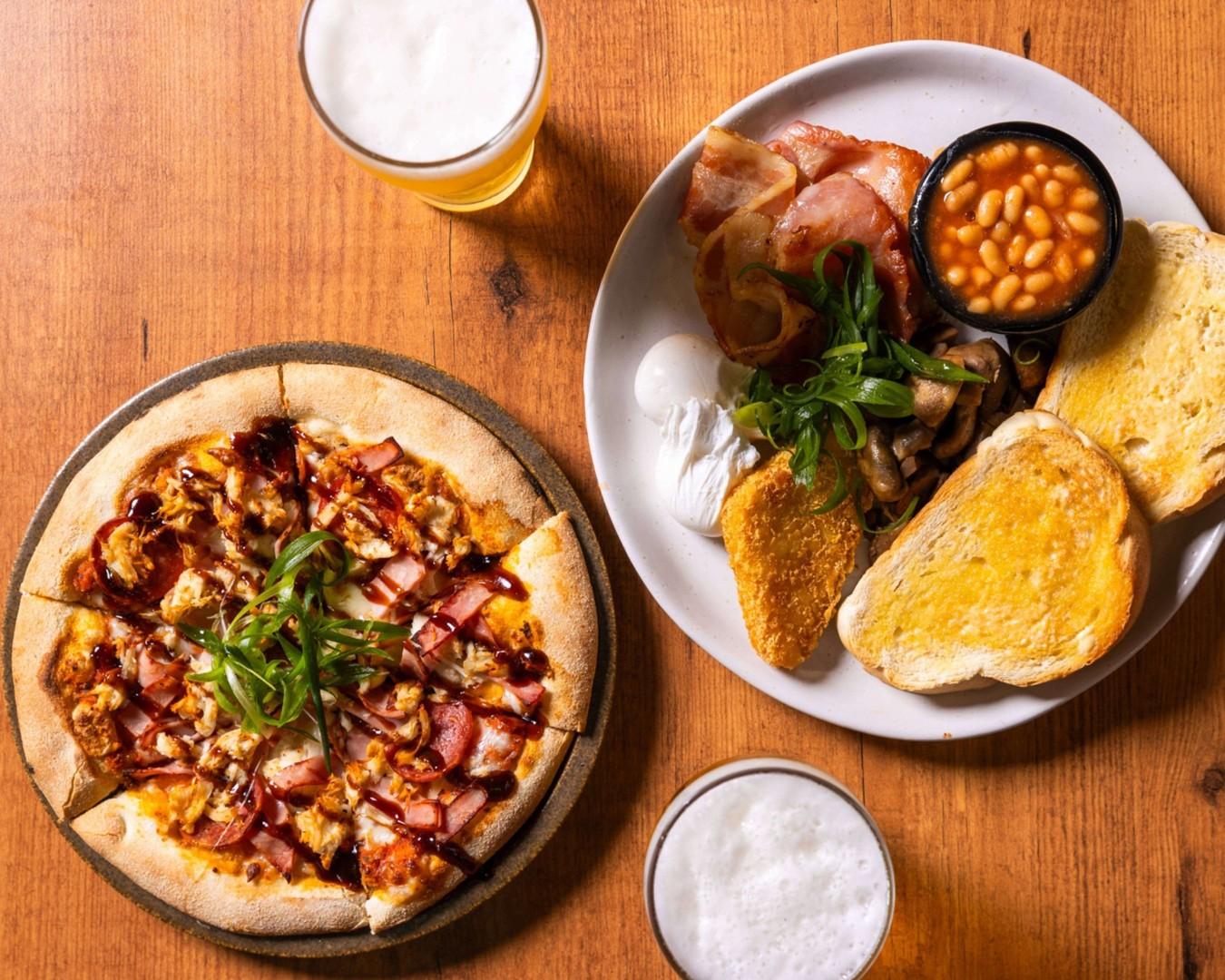 A wooden table at Townsville bar with a chicken and bacon barbecue pizza, a full breakfast plate, and two glasses of beer.