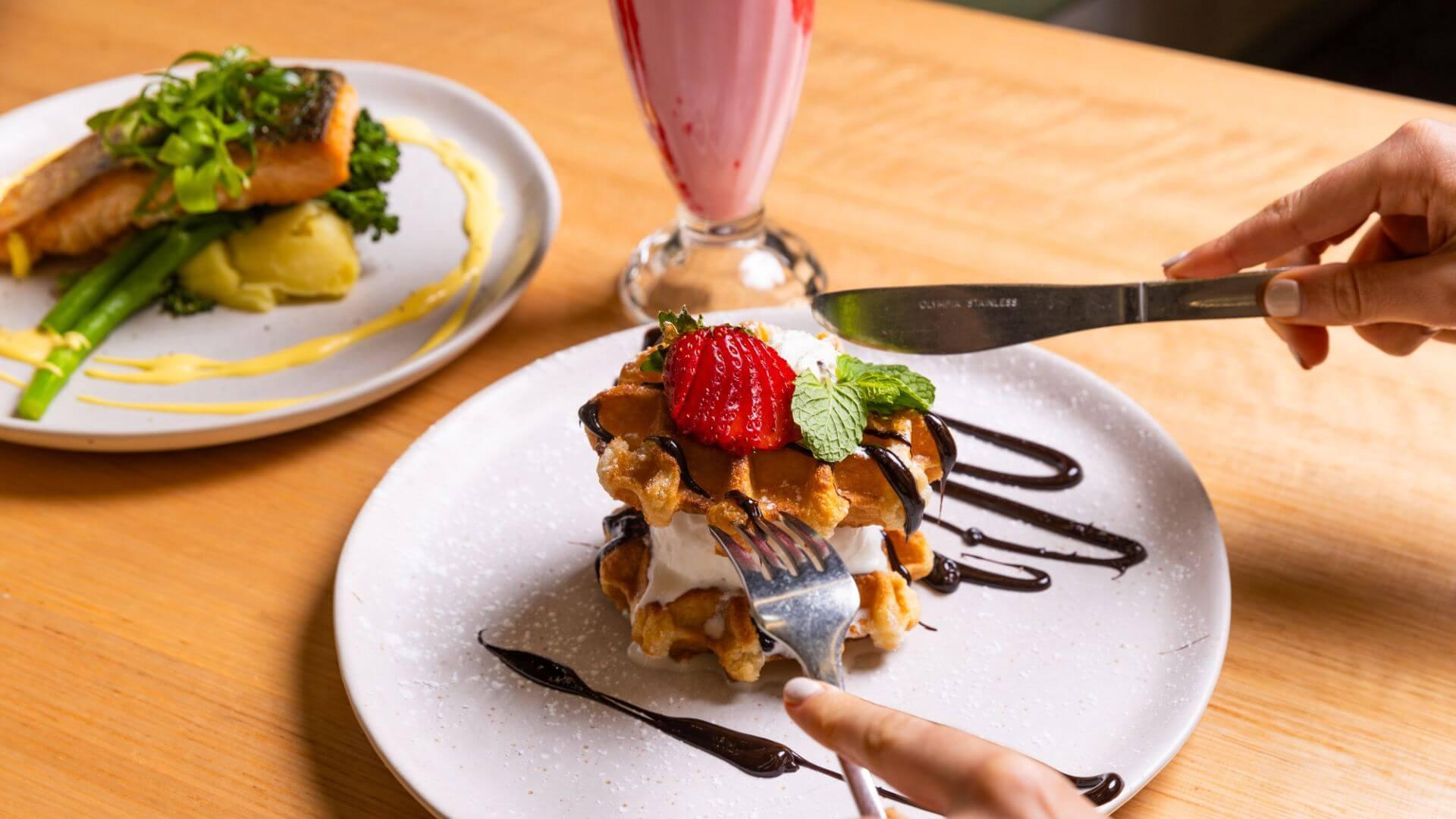 Close-up of waffles with whipped cream, strawberries, and chocolate sauce being cut with knife and fork, with salmon on mashed potatoes and a strawberry smoothie in the background.