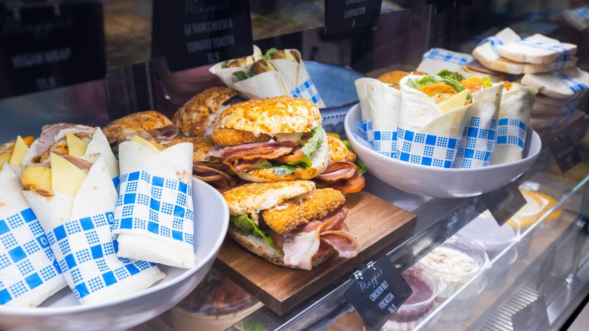 A display case at Maggie's Cafe filled with assorted sandwiches and wraps, including breaded chicken cutlet sandwiches and wraps in blue and white checkered paper.