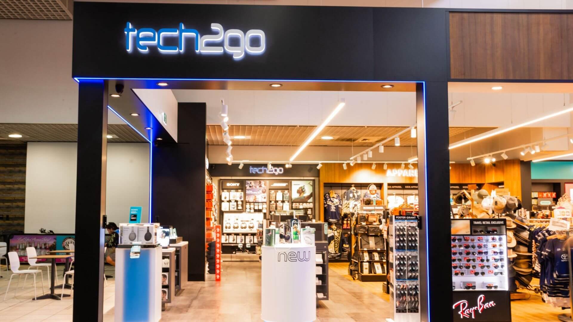 Entrance of tech2go store with headphones and gadgets on display under blue neon signage.