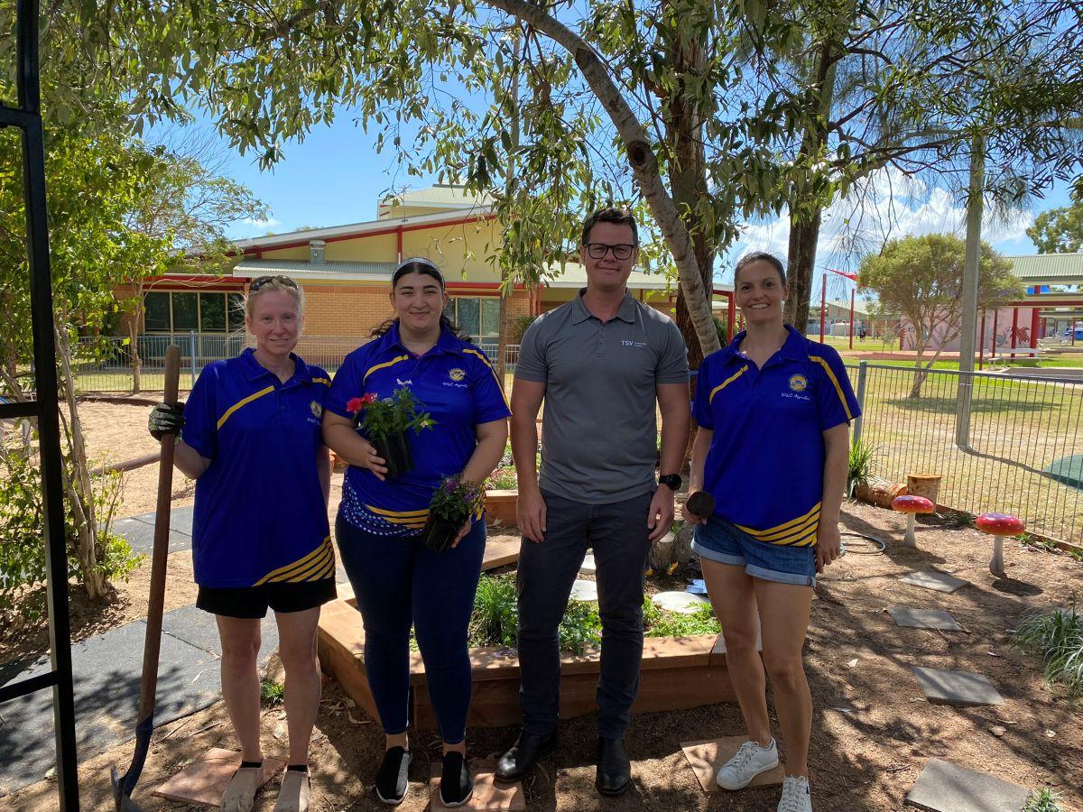 Four people standing in a garden area with trees, raised beds, and decorative mushrooms, including three in matching blue and yellow shirts and one in grey.