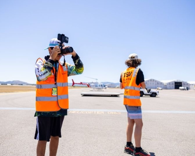 Townsville Airport photographers in high-visibility orange vests standing in the designated helicopter landing zone, with helicopters, airport infrastructure, and mountains visible in the backdrop.