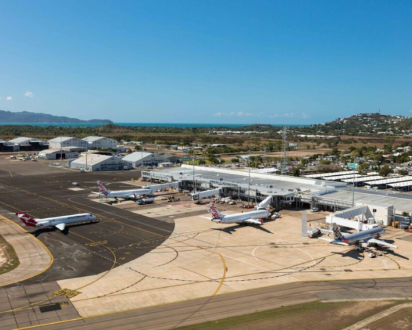 Aerial view of Townsville Airport showing terminals, hangars, and parked aircraft, with a runway on the left and coastal hills in the background.