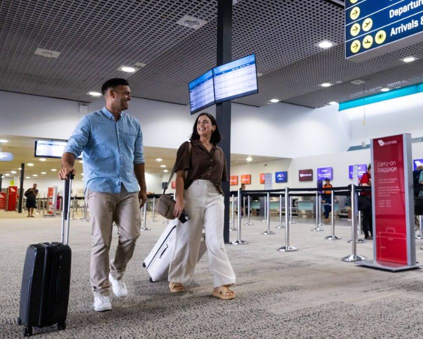 Passengers with luggage walk through a busy Townsville Airport check-in area, with multiple airline counters and digital flight information screens visible in the background. 