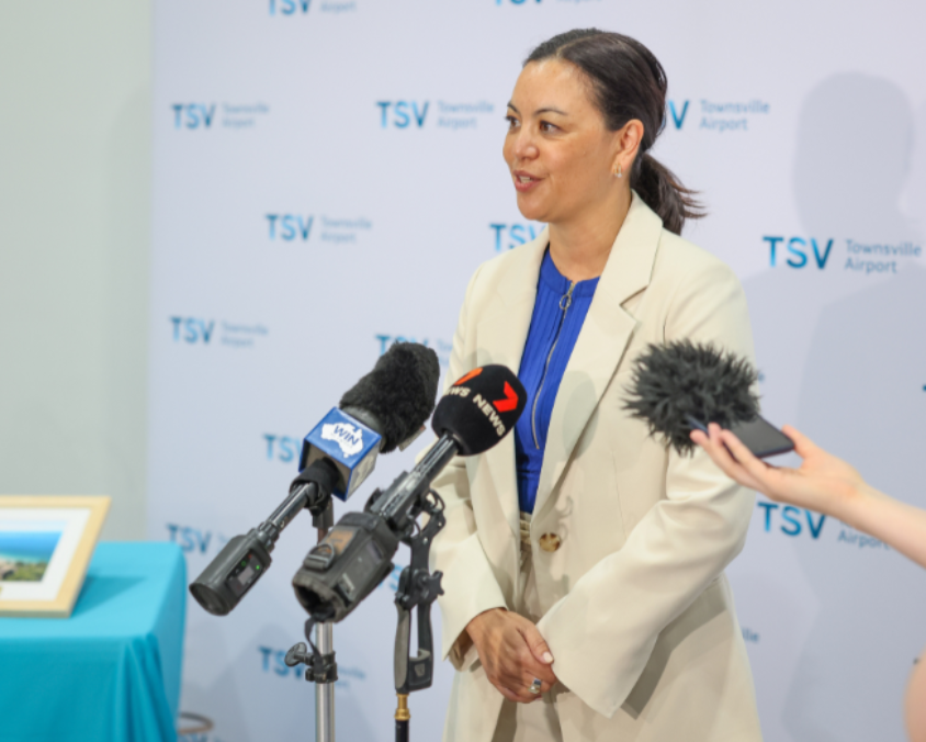 A senior spokesperson at Townsville Airport speaking to media outlets during a press event, with branded backdrop and multiple microphones.