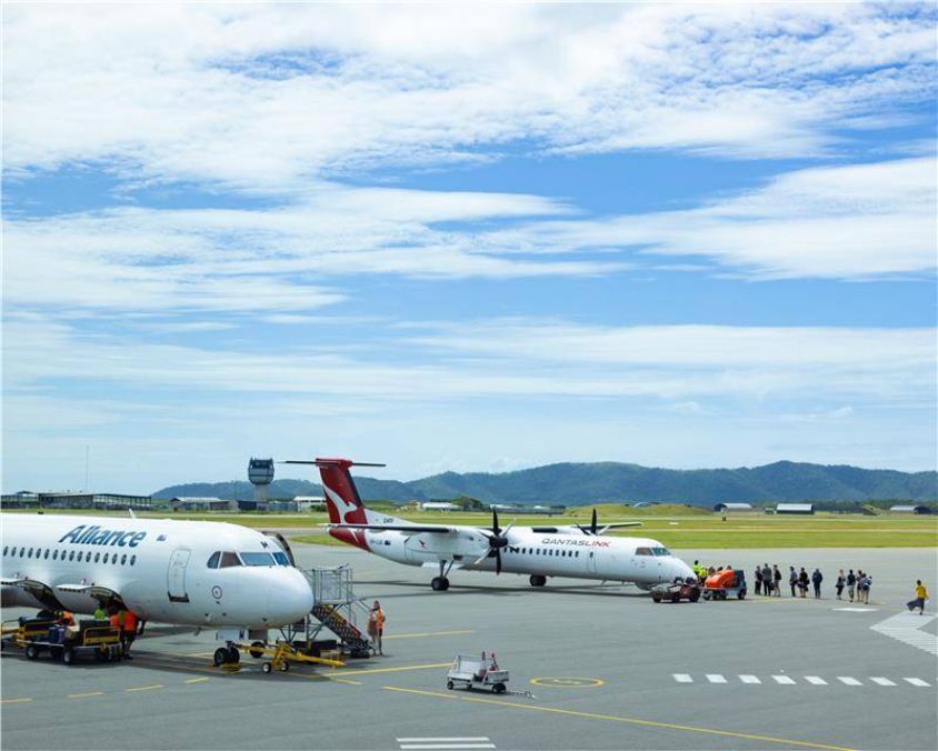 Alliance and QantasLink aircraft parked on Townsville Airport tarmac with ground crew and passengers nearby, control tower and hills in background.