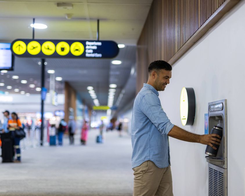 Traveler refills a water bottle at an airport hydration station, with directional signage and terminal activity visible in the background. 