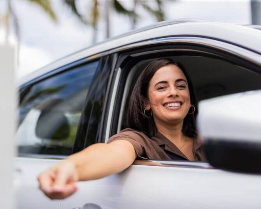 Smiling woman sitting in the driver’s seat of a car, leaning her arm out of the window while holding a parking ticket