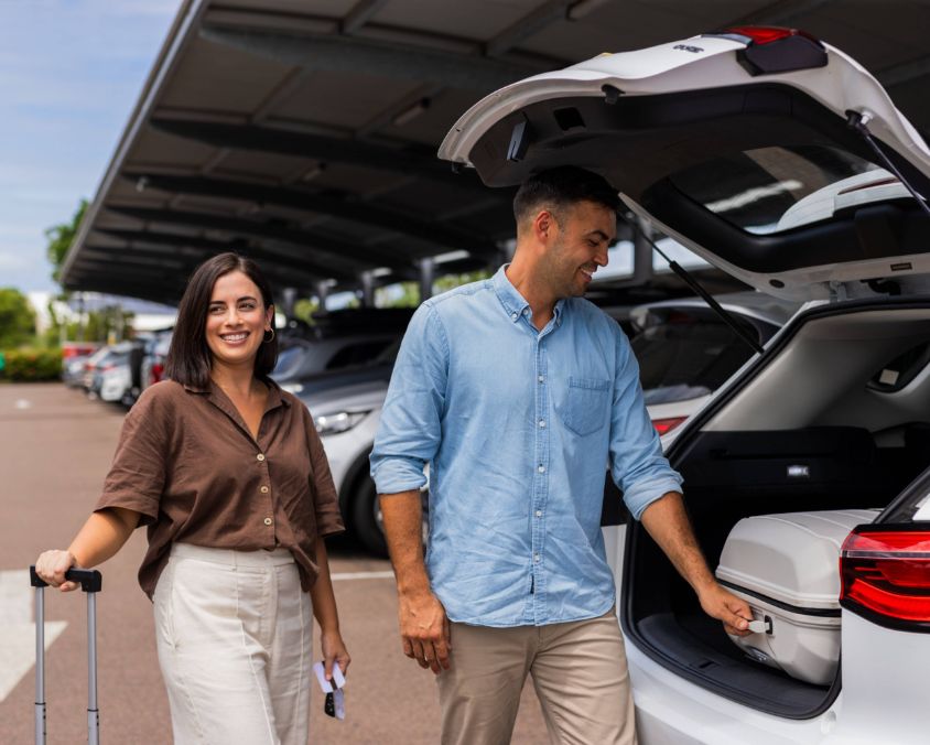 Two travelers in a covered airport parking lot, one handling luggage and documents while the other takes a suitcase out of a car trunk, surrounded by parked vehicles. 