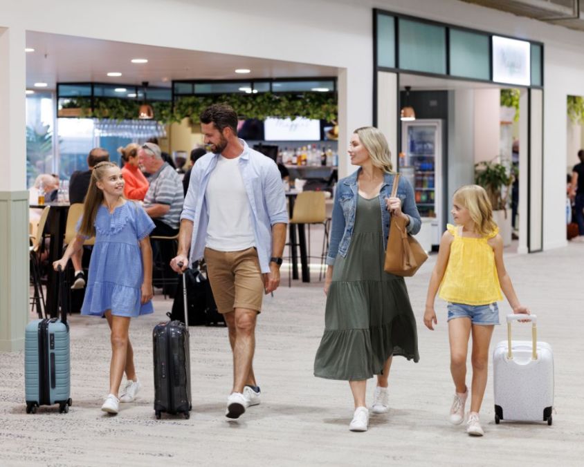 A family of four walking through the Townsville airport terminal with luggage, passing a dining area in the background.