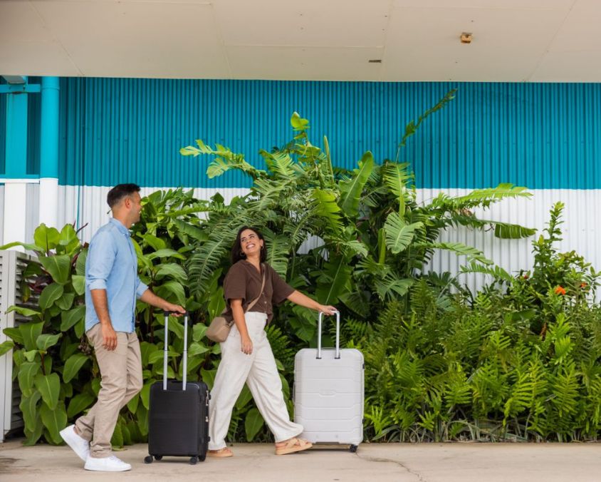 Two travelers walk with rolling suitcases in front of a blue and white building, surrounded by lush green plants. 