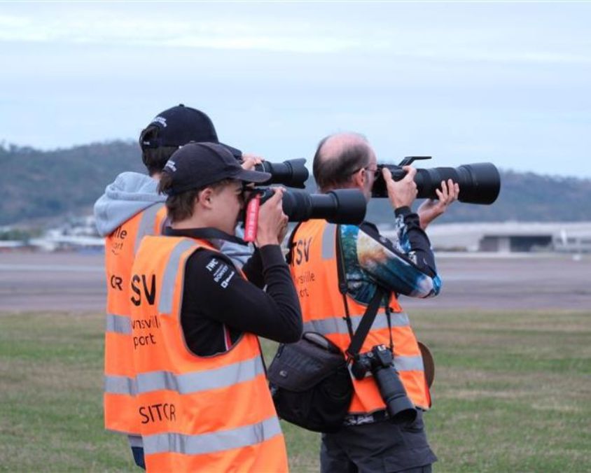 Photographers in high-vis vests marked “TSV Townsville Airport” near runway with cameras and distant hills.