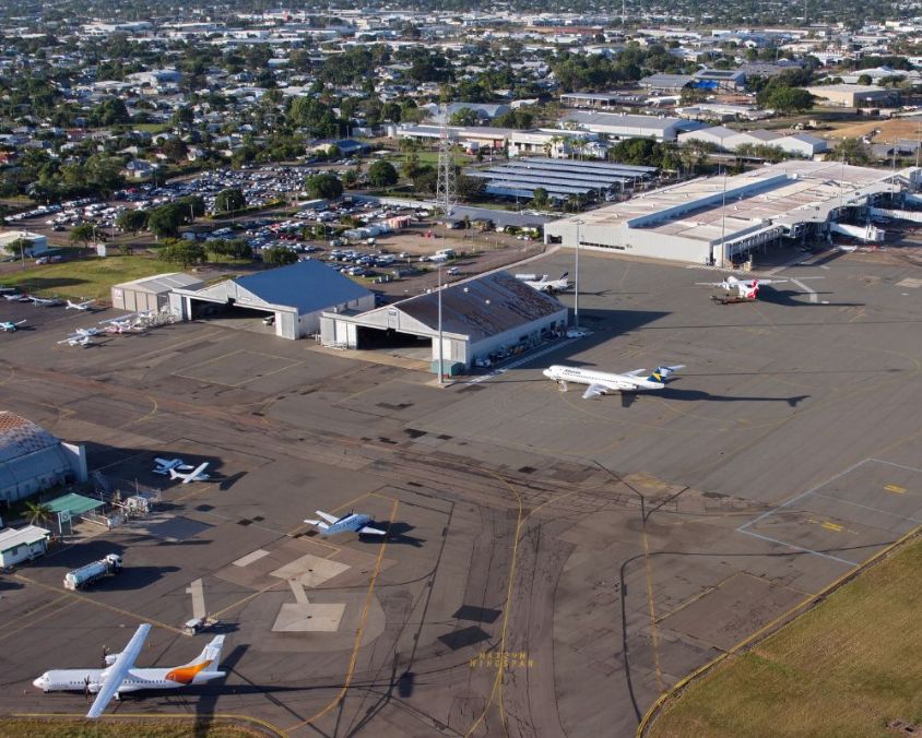 Aerial view of Townsville Airport with aircraft parked on the tarmac, multiple hangars and support buildings, a large parking area, and surrounding urban greenery.