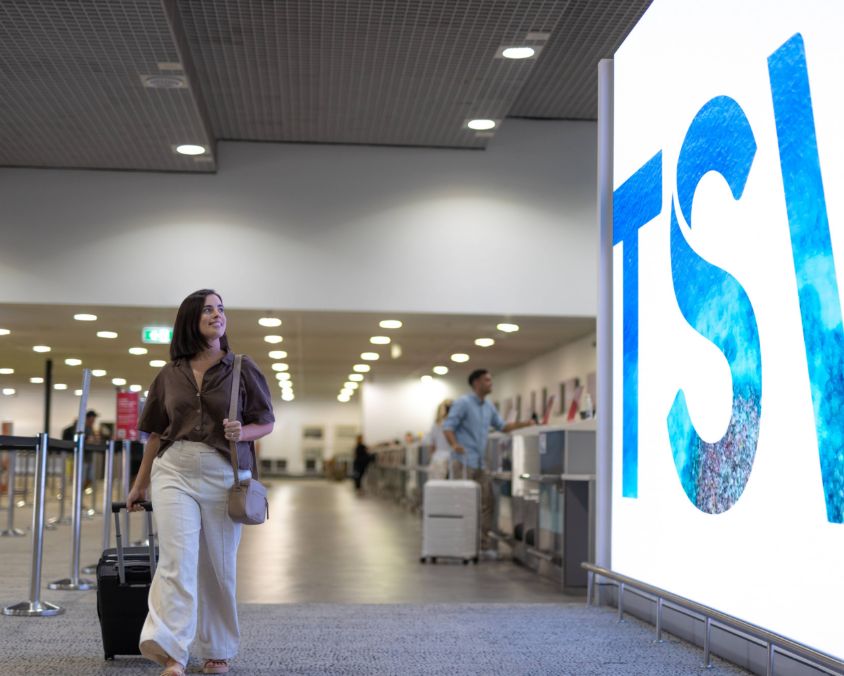 A traveler walks through Townsville airport terminal pulling a suitcase, with other passengers, check-in counters, and a large illuminated "TSV" sign in the background. 