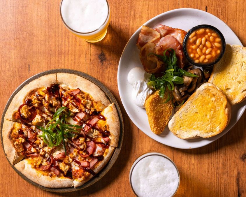 A wooden table at Townsville bar with a chicken and bacon barbecue pizza, a full breakfast plate, and two glasses of beer.