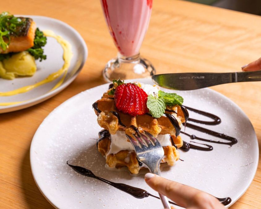 Close-up of waffles with whipped cream, strawberries, and chocolate sauce being cut with knife and fork, with salmon on mashed potatoes and a strawberry smoothie in the background.