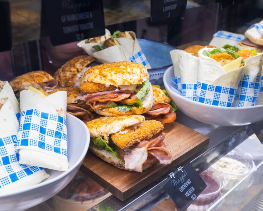 A display case at Maggie's Cafe filled with assorted sandwiches and wraps, including breaded chicken cutlet sandwiches and wraps in blue and white checkered paper.