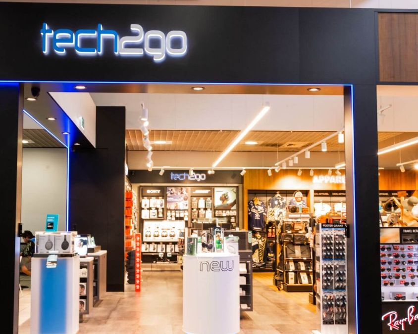 Entrance of tech2go store with headphones and gadgets on display under blue neon signage.