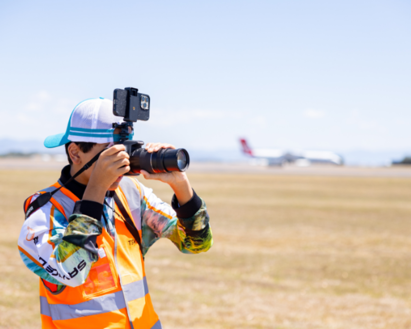 Townsville Plane Spotter's contestant in an orange safety vest taking a photograph on the airfield, with an airplane visible in the background.
