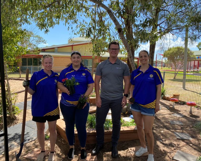 Four people standing in a garden area with trees, raised beds, and decorative mushrooms, including three in matching blue and yellow shirts and one in grey.