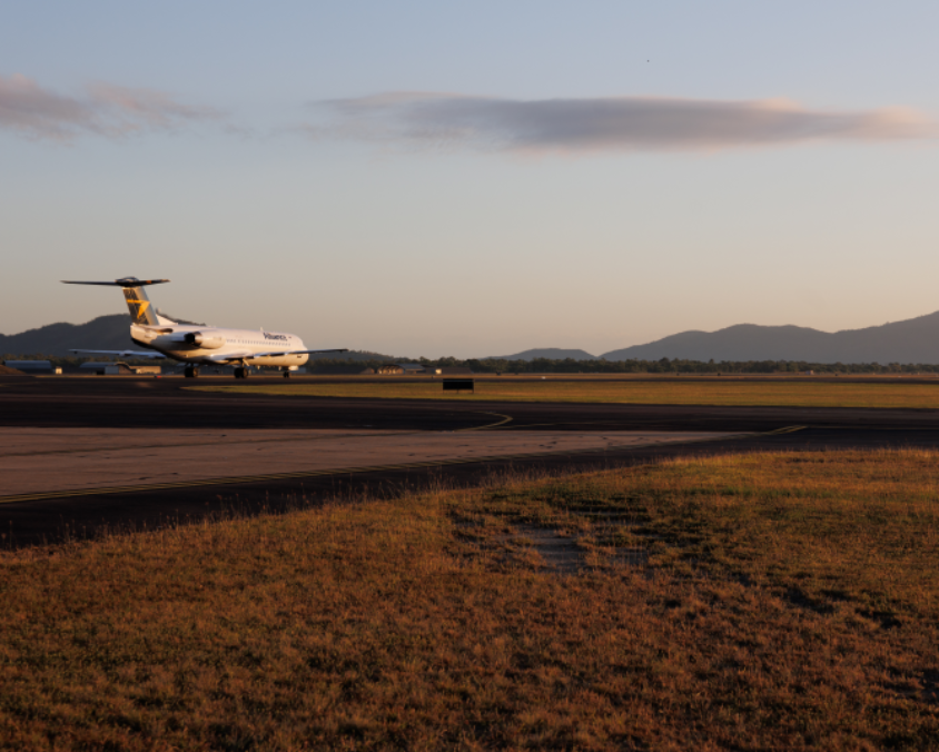 An Alliance Airlines McDonnell Douglas MD-80 series aircraft on the Townsville runway during sunrise or sunset, with rear-mounted engines and a T-tail, set against a mountainous backdrop.