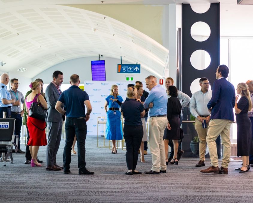 Community consultation group meeting inside Townsville Airport terminal, with participants conversing beneath curved ceiling panels and directional signage.