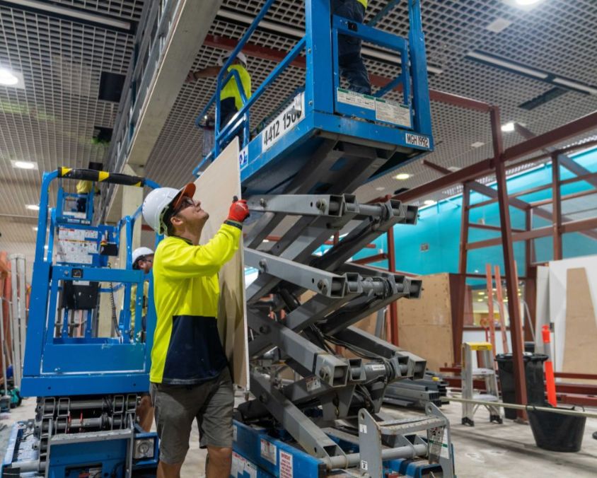 Construction workers using a scissor lift at Townsville Airport, with one holding a large panel and another on the elevated platform, surrounded by tools and materials.