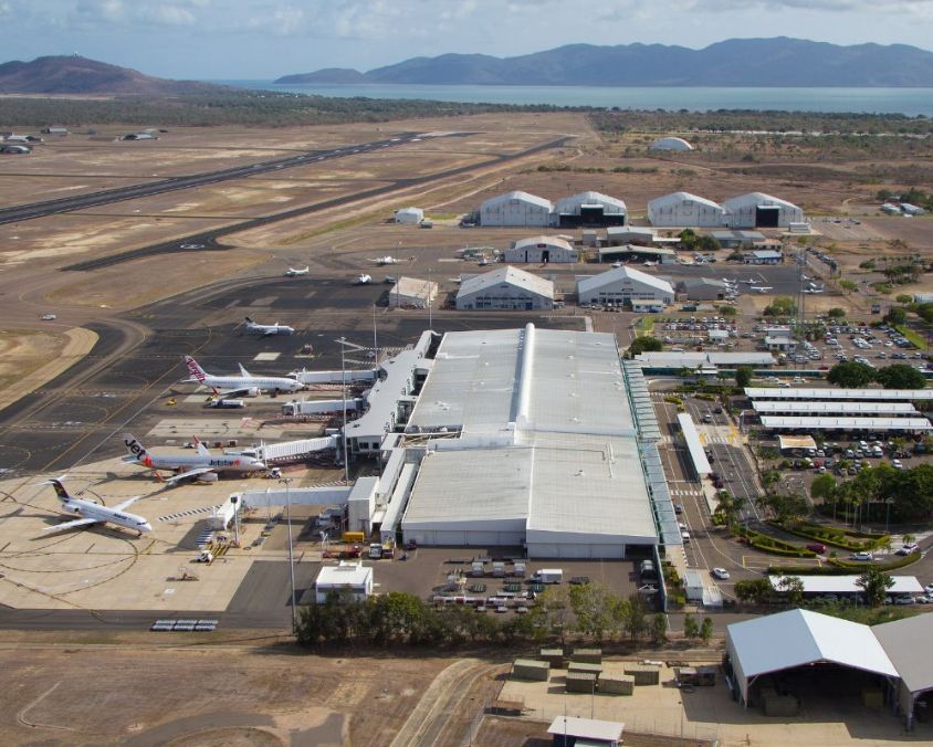 Aerial view of Townsville airport with planes at gates, hangars, runways, and surrounding roads near mountains and water.