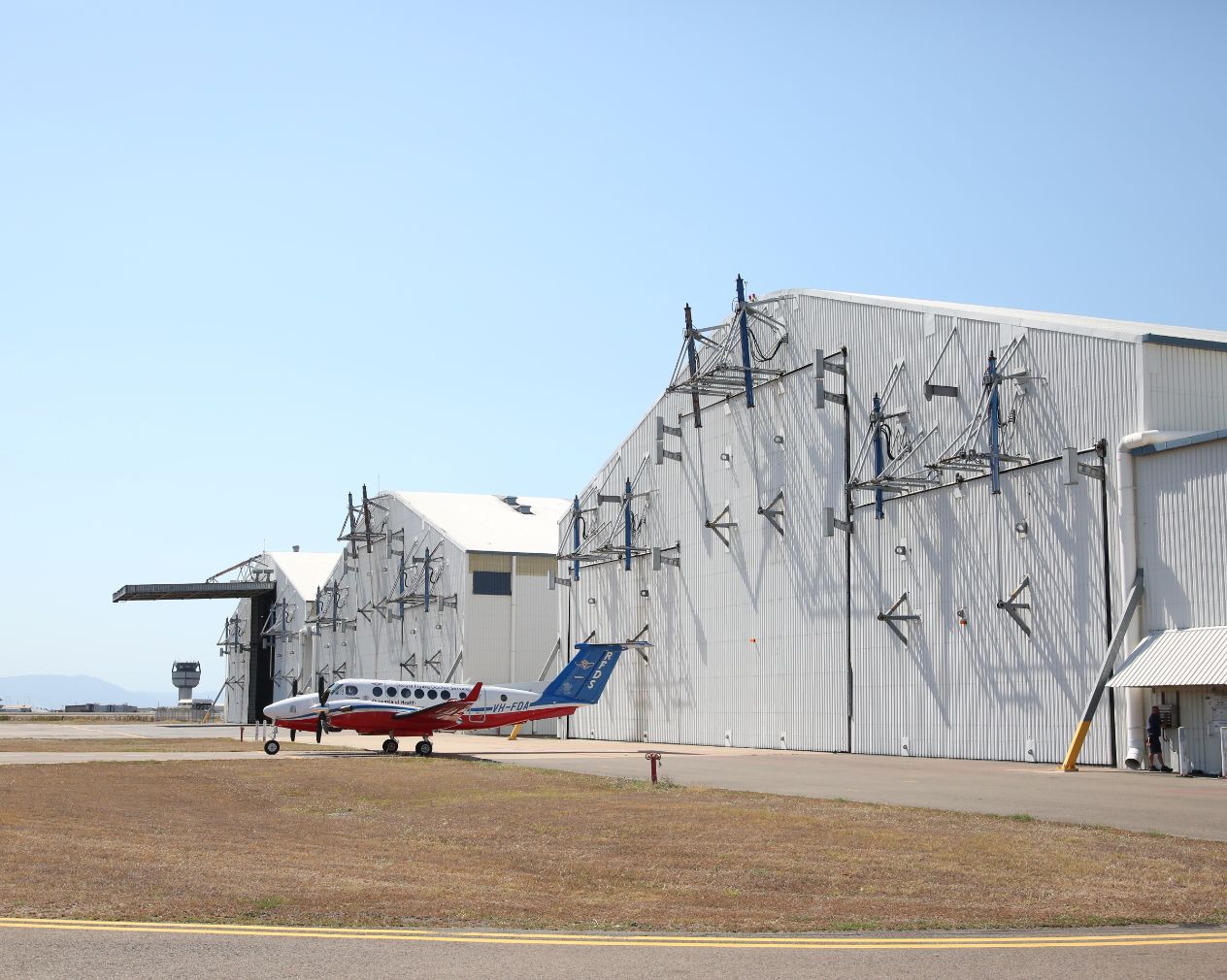 Small aircraft parked on the tarmac near a multi-section hangar at Townsville Airport, with a control tower in the background and clear skies overhead.