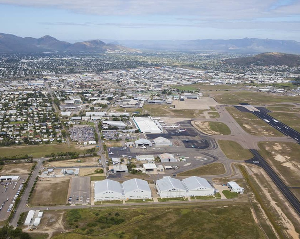 Aerial view of Townsville Airport showing white-roofed terminal buildings, adjacent runway, and surrounding urban and mountainous landscape.