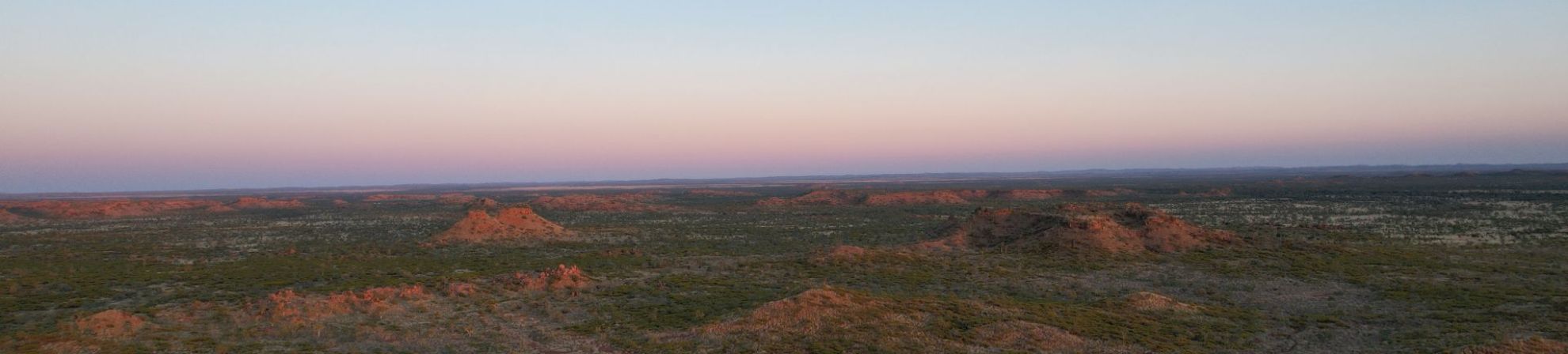 Wide view of an arid landscape with scattered hills under a clear sky transitioning from blue to pink near the horizon.