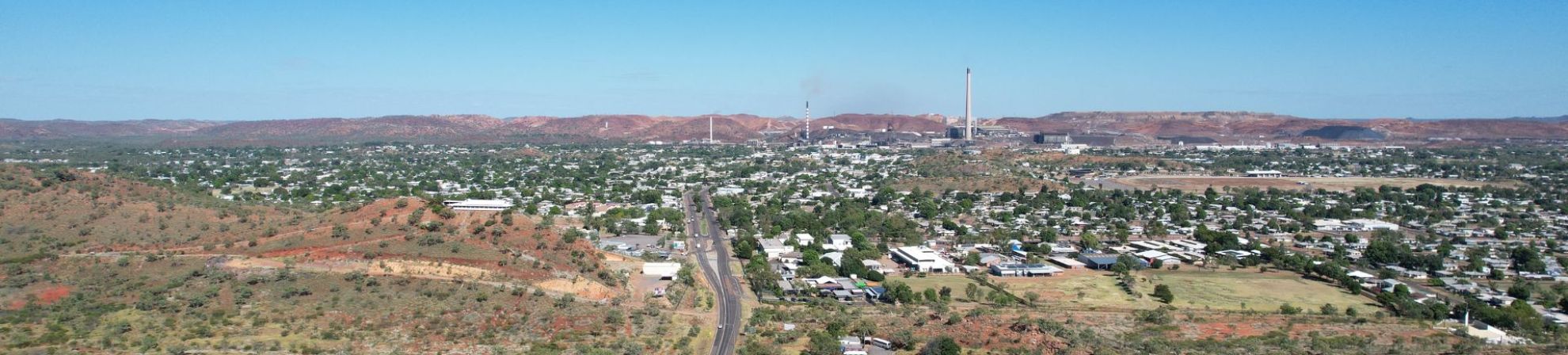 An aerial view of Mount Isa town with residential and industrial areas, a central road leading to smokestacks, surrounded by hilly terrain and red-colored hills under a clear blue sky.